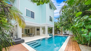 A two-story house with a swimming pool surrounded by wooden decking and tropical plants.