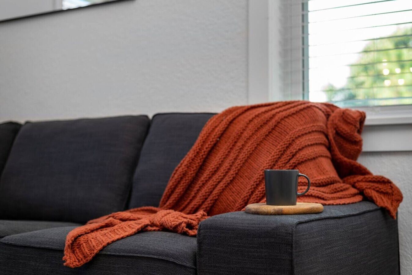 Un sofá gris oscuro con una manta naranja de punto y una taza oscura sobre un posavasos de madera.