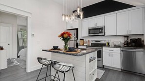 A modern kitchen with white cabinetry, a central island with stools, a hanging light fixture, and stainless steel appliances. - The BoHo Bungalow - Updated Space Close To Beach! (Kill Devil Hills)