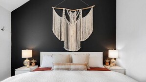 A bed with a white bedspread, two bedside lamps, and a macramé wall hanging. - The BoHo Bungalow - Updated Space Close To Beach! (Kill Devil Hills)