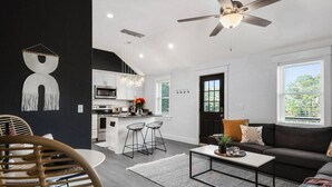 A modern living room with a dark accent wall, a large window, a ceiling fan, and a kitchen area with white cabinets and a central island. - The BoHo Bungalow - Updated Space Close To Beach! (Kill Devil Hills)