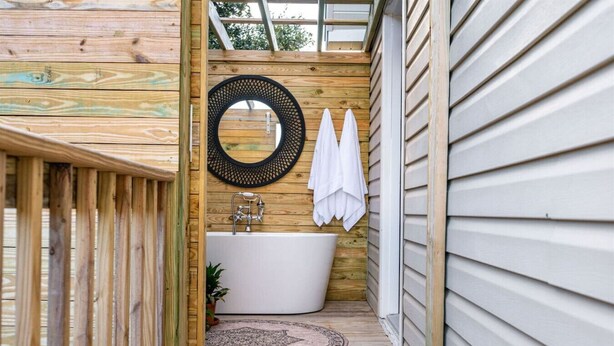 A wooden deck leading to a bathroom with a freestanding bathtub, a round mirror, and two white towels hanging on the wall. - The BoHo Bungalow - Updated Space Close To Beach! (Kill Devil Hills)