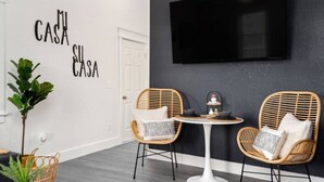 A modern room with two wicker chairs, a small round table, a potted plant, and a wall-mounted TV. - The BoHo Bungalow - Updated Space Close To Beach! (Kill Devil Hills)