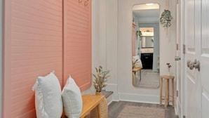 Un couloir avec une armoire rose, un miroir, une petite table avec un vase et un banc avec des coussins.
