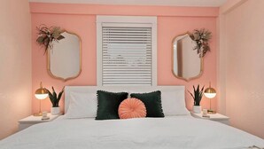 Une chambre à coucher comprenant un lit, deux tables de chevet, une fenêtre avec des stores, deux miroirs muraux et un luminaire au plafond.
