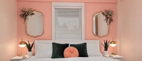 Un dormitorio con una cama, dos mesitas de noche, una ventana con persianas, dos espejos en la pared y una lámpara en el techo.