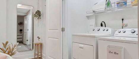 A laundry room with a front-loading washer and dryer, a small stool, and a shelf with cleaning supplies.