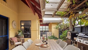 Een terras buiten met een houten tafel, rieten stoelen, een barbecue en uitzicht op de achtertuin.