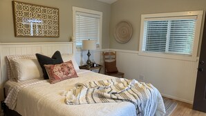 A bedroom with a bed, pillows, a bedside table, a lamp, a chair, and two windows with blinds. - Cottage studio (Poway)