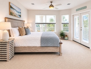 A bedroom with a bed, two bedside lamps, a ceiling fan, and a view of the outdoors through large windows.