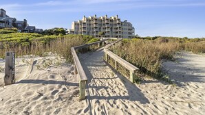 Un paseo marítimo de madera que lleva a un edificio frente a la playa con varios balcones y una playa arenosa con vegetación densa.
