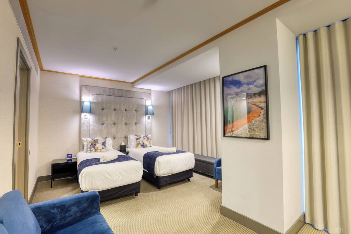 A hotel room with two beds, a blue armchair, a nightstand, a wall-mounted lamp, and a framed picture.