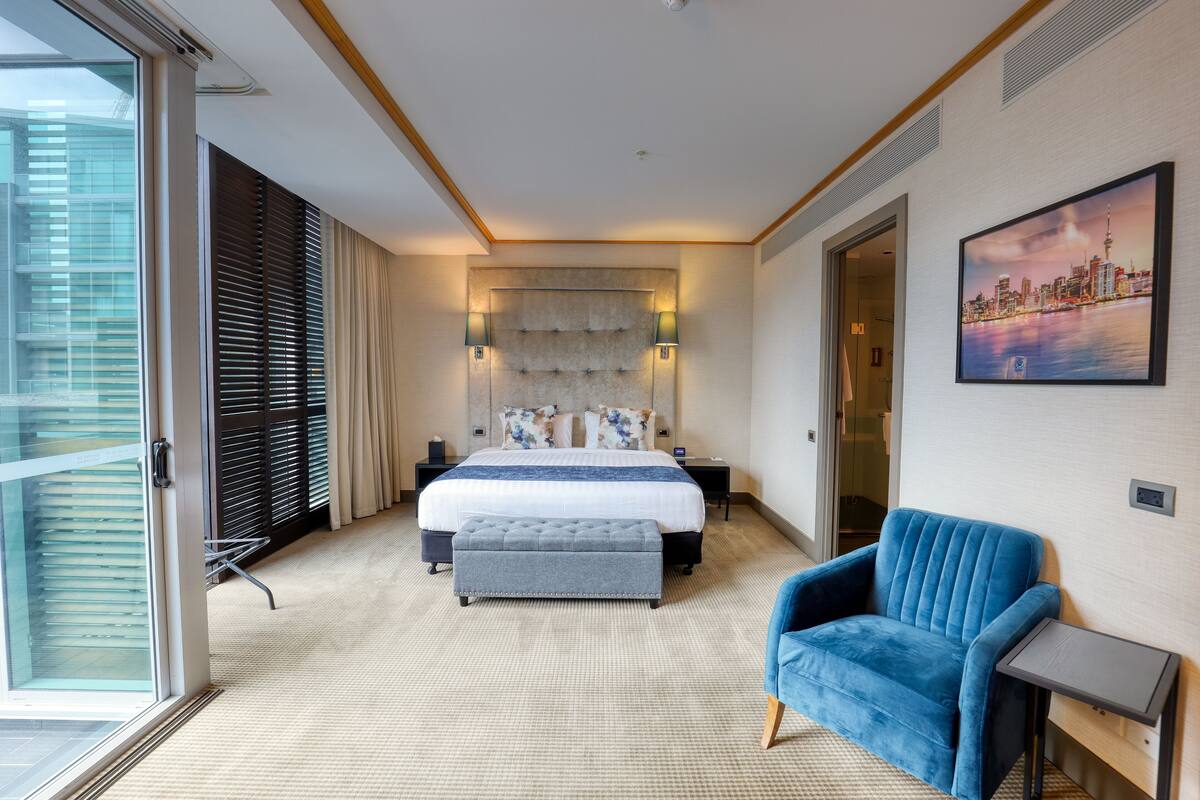 A hotel room with a large bed, a blue armchair, a small table, and a cityscape painting on the wall.