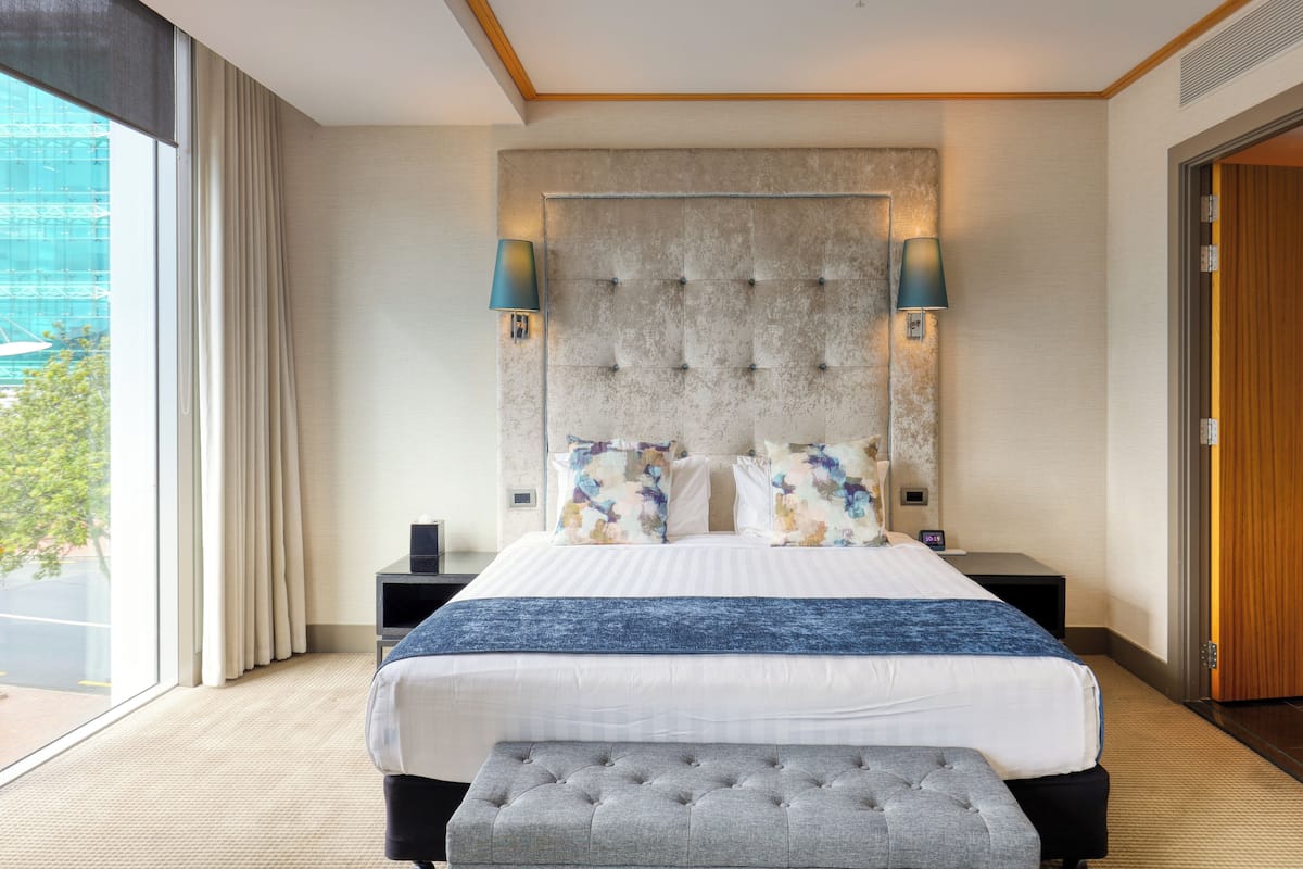 A hotel room with a large bed, a headboard, two bedside lamps, a nightstand, and a view of the outdoors.