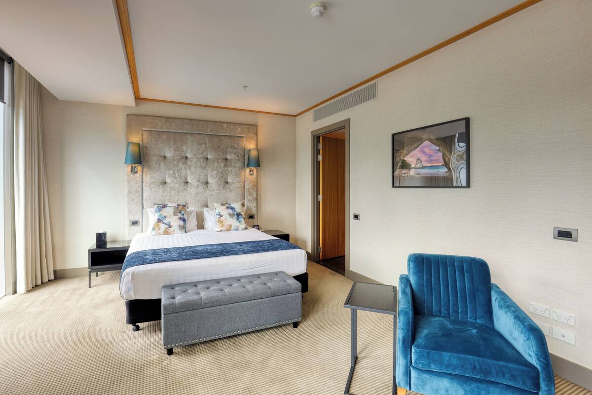 A hotel room with a large bed, a grey ottoman, a blue armchair, a small table, and a painting on the wall.