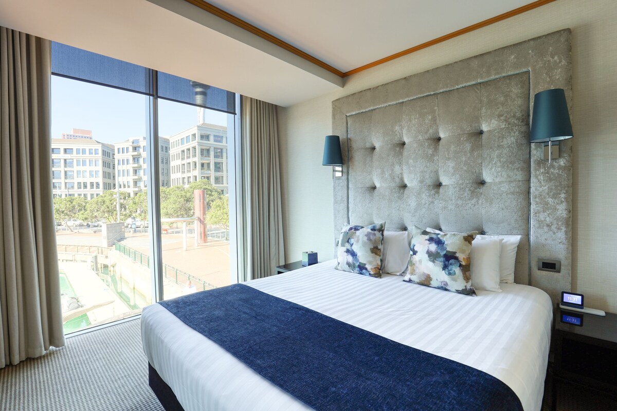 A hotel room with a large bed, a view of buildings through the window, and a modern headboard.