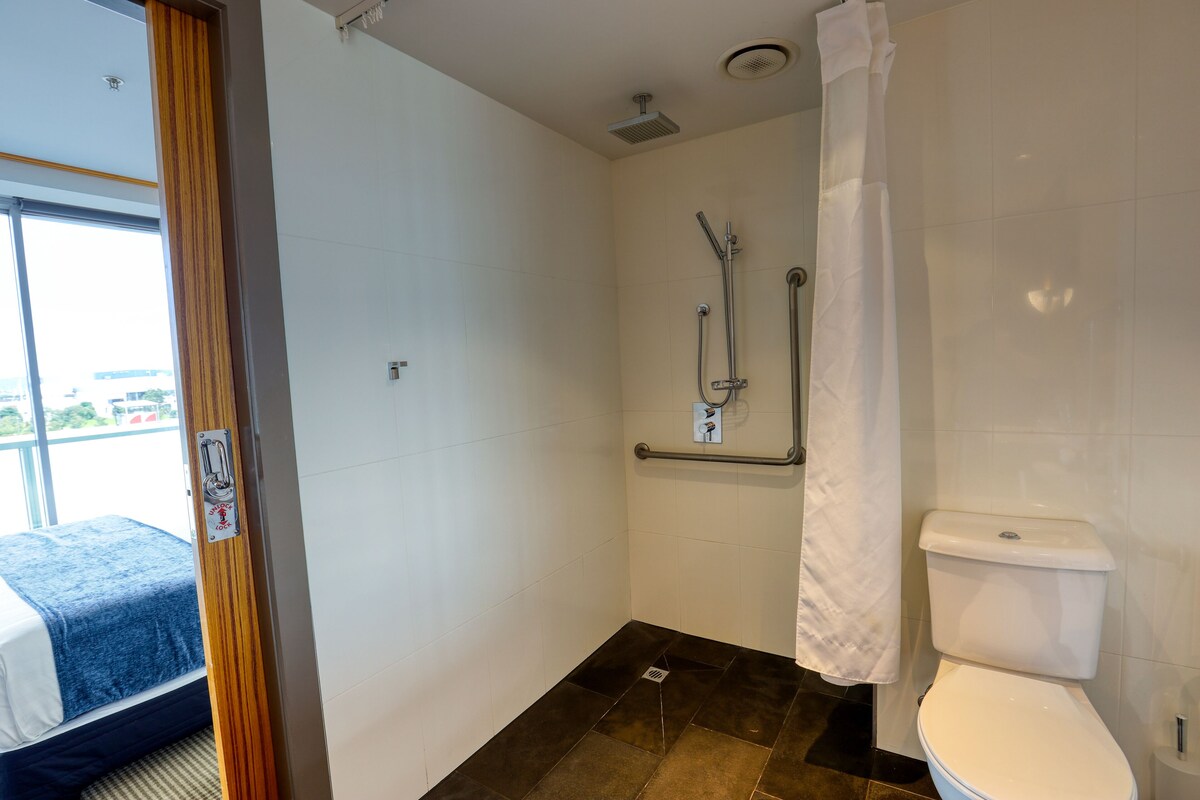 A bathroom with a shower, toilet, and a bed visible through an open door.