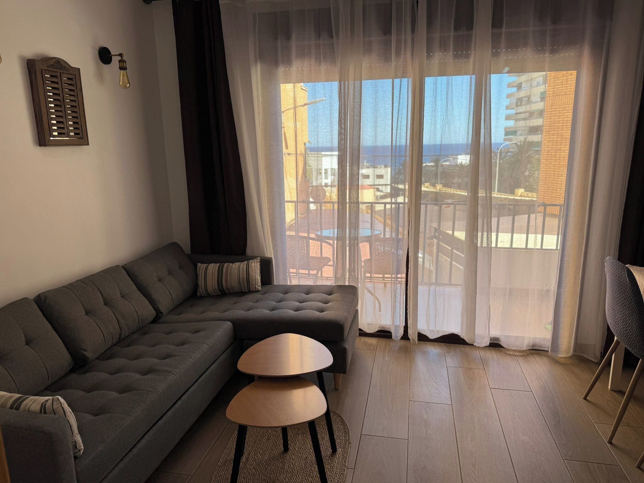Una sala moderna con un sofá seccional gris, dos mesitas redondas, cortinas translúcidas y vista a la ciudad a través de amplias ventanas.