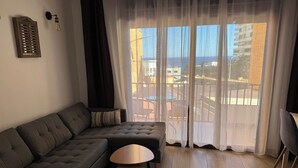 A modern living room with a grey sectional sofa, two small round tables, sheer curtains, and a view of the cityscape through large windows. - Sunny Seaside Apartment in Aguadulce (AGUADULCE)