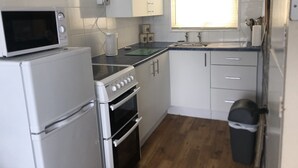 Une cuisine de taille réduite, avec des appareils électroménagers blancs, une fenêtre avec des stores et une poubelle.
