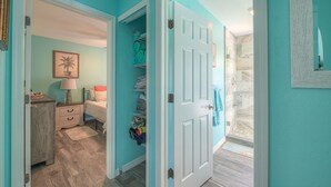 Un couloir avec un mur de couleur turquoise, une porte blanche et un plancher en bois. On y trouve un lit avec une table de chevet et une lampe, ainsi qu’une armoire contenant des vêtements pliés.
