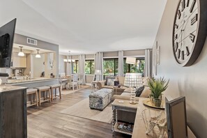 Amplia sala de estar con cocina, un reloj de pared grande y un comedor con taburetes de bar.