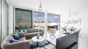 A modern living room with a sofa, coffee table, and TV, a dining area with chairs and a table, and large windows with a view of buildings and sky. - Villa Prol26, Stunning 3bdr Protaras Villa with Pool, Close to Fig Tree Bay (Protaras)