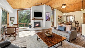 A spacious living room with a fireplace, wooden ceiling, and large windows offering a view of the outdoors.