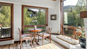 A room with large windows, a wooden ceiling, a round table with chairs, a bench, and a potted plant.