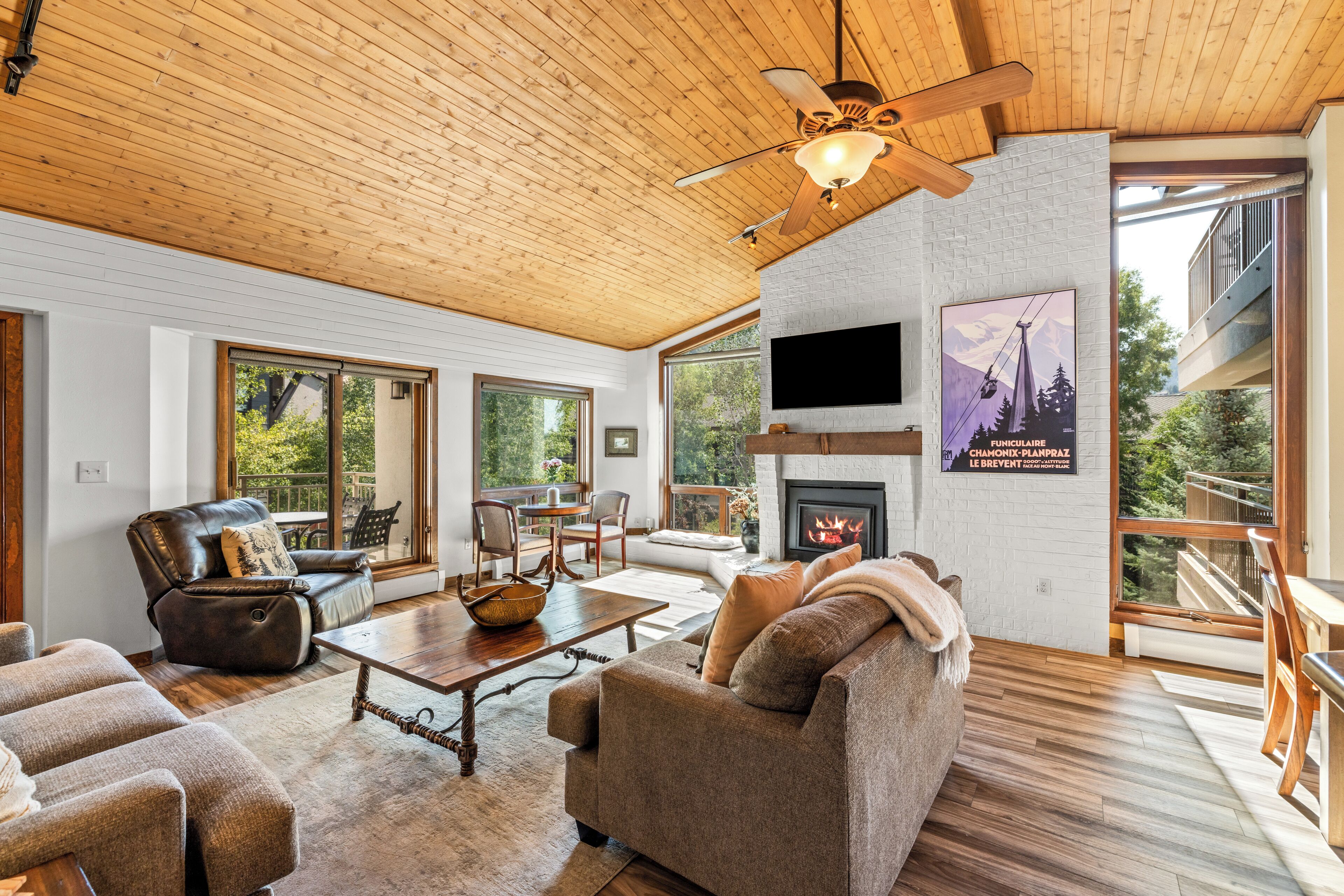 A spacious living room with a fireplace, wooden ceiling, and large windows offering a view of the outdoors.
