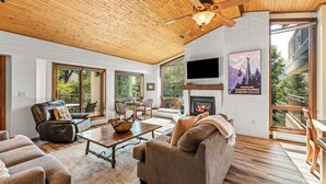 A spacious living room with a fireplace, wooden ceiling, and large windows offering a view of the outdoors.