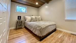 A bedroom with a wooden ceiling, a bed with a white and gray duvet, a small table with a sign, and a window with blinds. - Eastside Modern Farmhouse in downtown Kalispell (Kalispell)