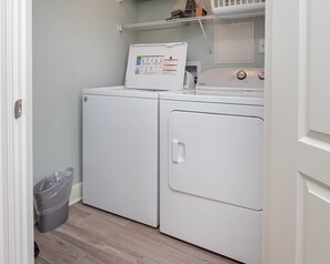 A laundry room with a front-loading washer and dryer, a trash can, and a shelf above the dryer.
