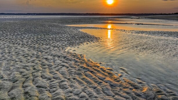 Une plage où l’on voit des empreintes de pas menant à l’eau, un coucher de soleil en arrière-plan et une mer calme.