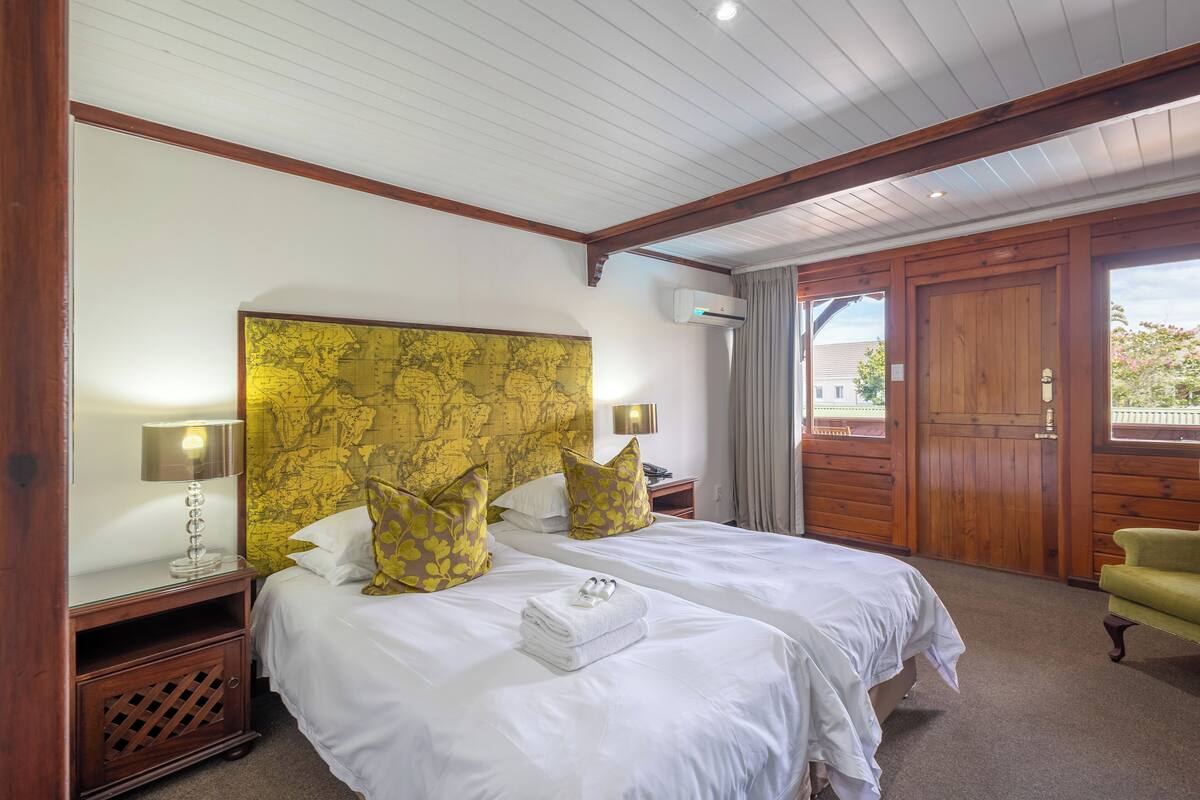 Habitación de hotel con dos camas, cabecero de madera, ropa de cama blanca, una mesita con una lámpara y vistas al exterior.