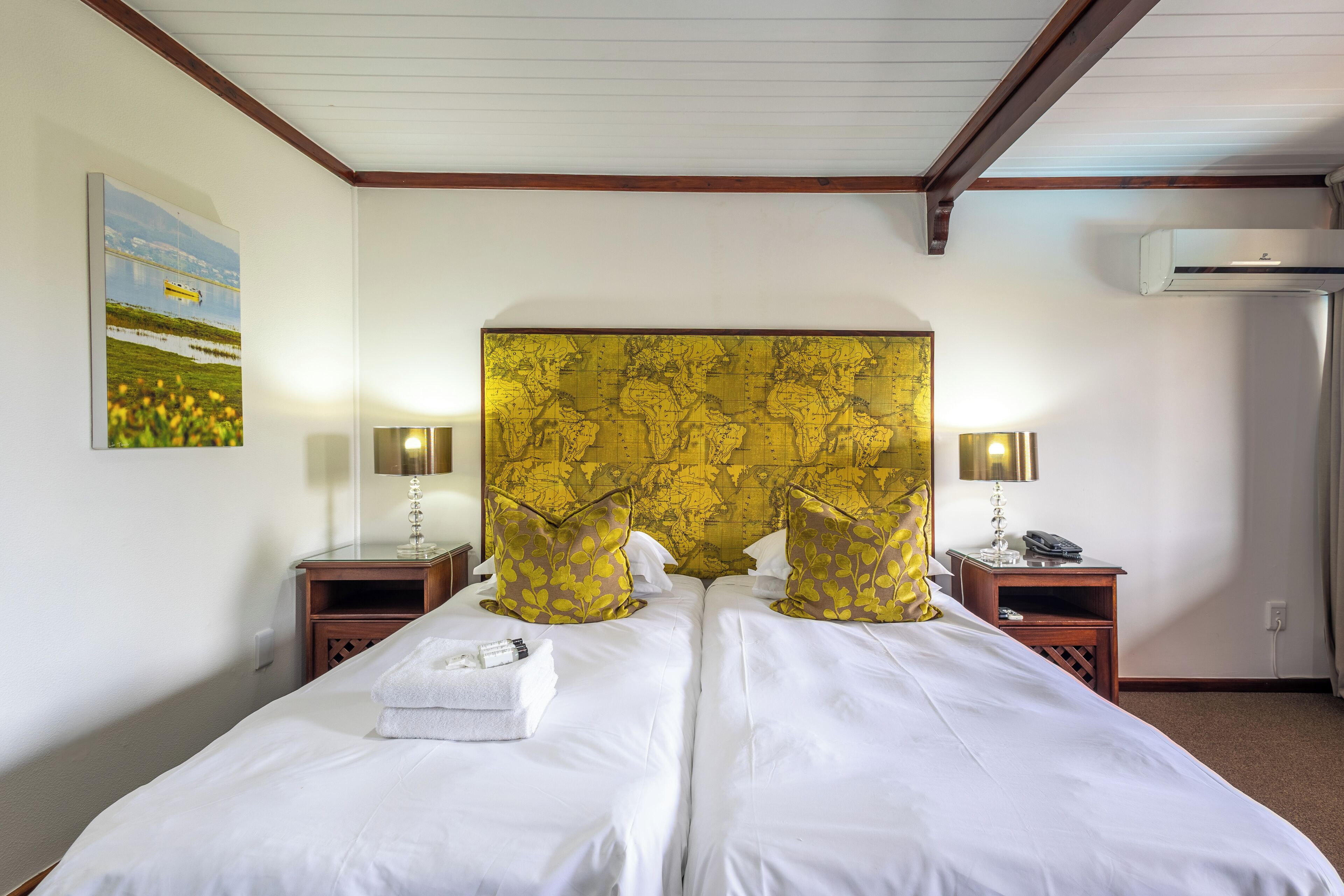 A double bed with white linens and gold-patterned headboard, two bedside tables with lamps, and a wall-mounted air conditioner.