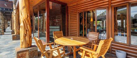 Una terraza exterior de madera con una mesa redonda y sillas, pilares de piedra y grandes puertas de cristal que dan al interior.