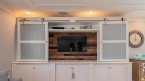 A modern living room with a built-in TV, wooden panel wall, white cabinetry, and a clock on the wall. - The Sassy Seahorse: Spacious Beach Home with Multiple Living Areas and Room for (Oak Island)