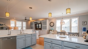 A modern kitchen with light blue cabinets, a central island, pendant lights, and a dining area with a table and chairs. - The Sassy Seahorse: Spacious Beach Home with Multiple Living Areas and Room for (Oak Island)