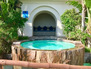 Una piscina circular con borde de madera, rodeada de vegetación y un edificio blanco con entrada arqueada.