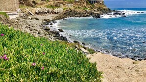 Une propriété située sur la côte, avec un rivage rocheux, une eau d’un bleu limpide et une vue sur un bâtiment blanc doté de nombreuses fenêtres et d’une tour.