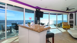 Une cuisine moderne dotée d’un grand îlot central, d’un coin repas avec une table en verre et d’une vue sur l’océan.