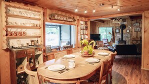 A wooden dining area with a round table, chairs, and a bookshelf filled with books and glassware. - Riverside Mid-Century Cabin with Mountain Views (Baring)