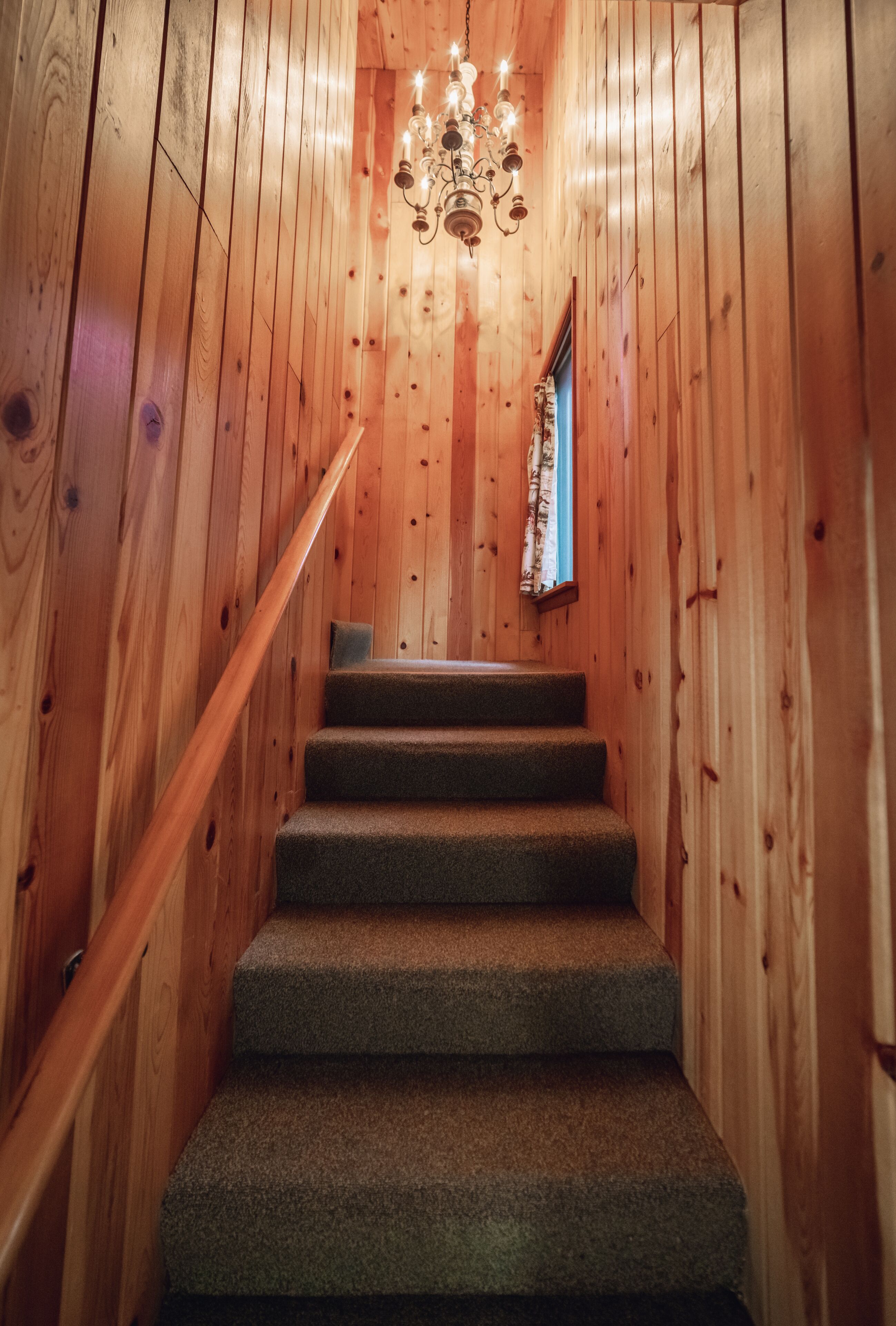A wooden staircase with carpeted steps leading to a room with a window and a chandelier.