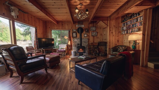 A cozy cabin interior with wooden walls, a fireplace, a television, leather furniture, and a bookshelf. - Riverside Mid-Century Cabin with Mountain Views (Baring)