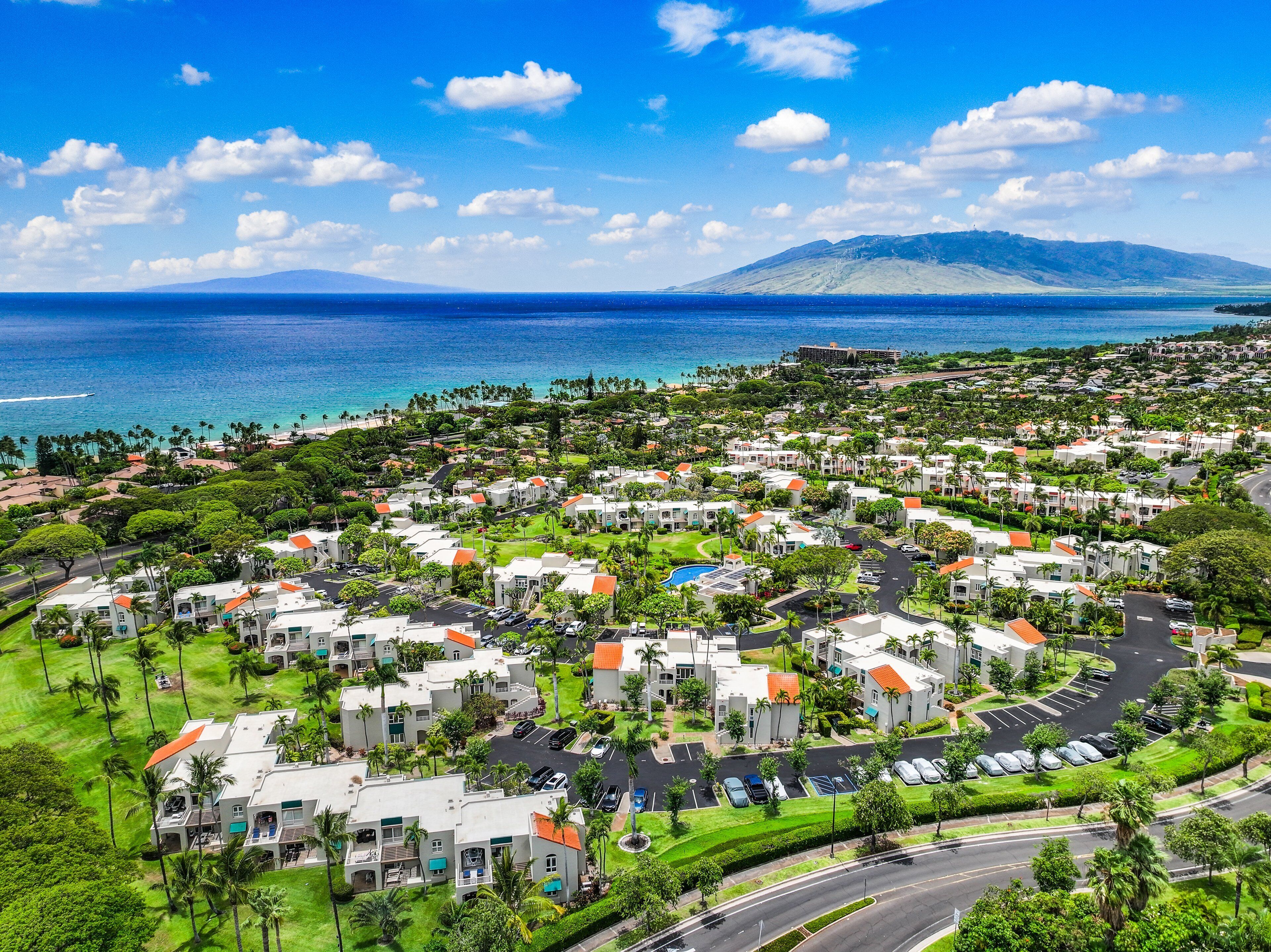 A coastal resort with multiple-story buildings, a central pool, and a view of the ocean and a mountain in the distance.