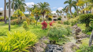 Un exuberante jardín con palmeras, una pequeña cascada y un edificio al fondo.