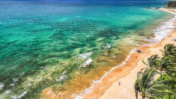Una playa con agua turquesa cristalina, una orilla arenosa y una montaña al fondo.