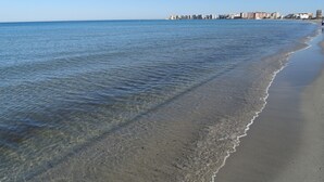 A beach with clear water, a sandy shore, and a cityscape in the background. - Duplex Aldeas Taray Club 55 Admer (La Manga del Mar Menor)