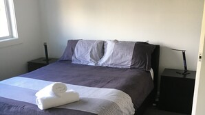 A neatly made bed with a dark-colored comforter and pillows, a folded white towel, and two bedside tables with lamps. - Entire House 3 bedrooms, 5 minute walk to town centre and 1 minute to beach (Strahan)